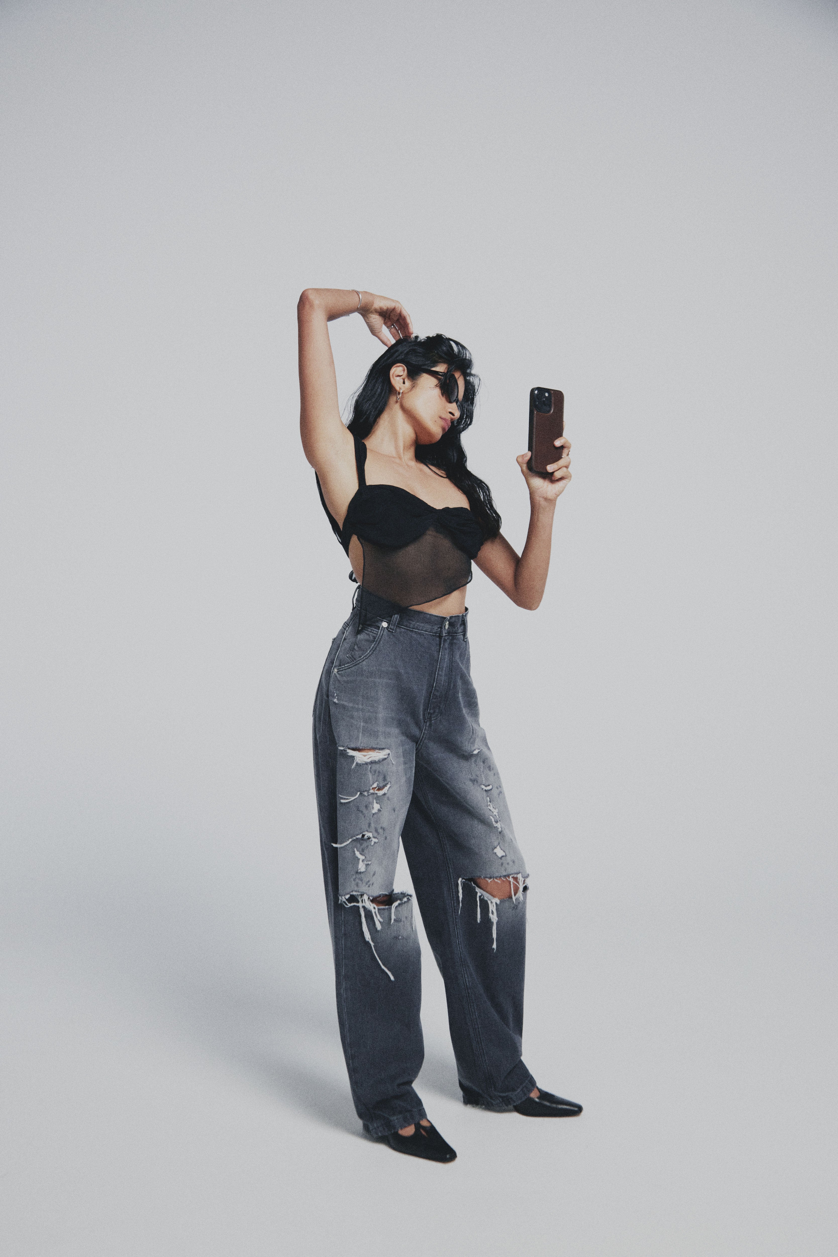 Woman wearing a black top and ripped jeans, taking a selfie on a gray background